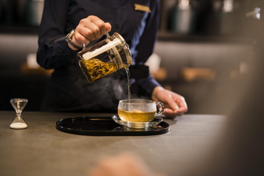A server pouring hot loose leaf tea into a large glass tea cup. Beside it is a small sand timer.